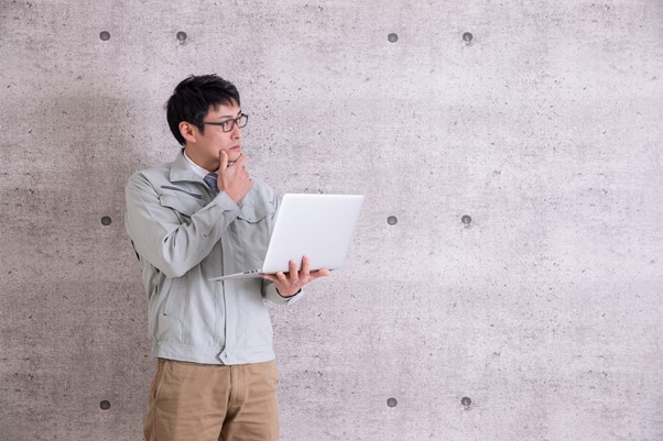 PCを持って考え込む作業着姿の男性